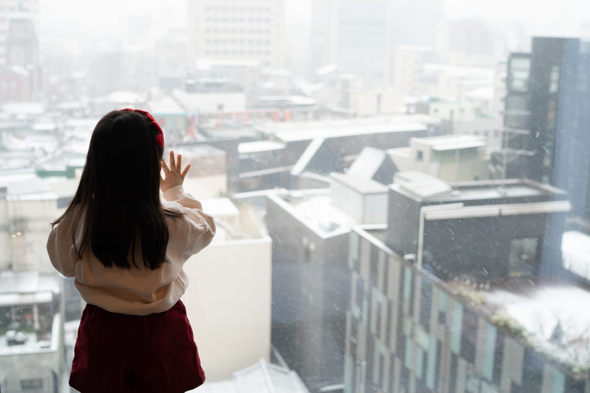 A child looks out through a window toward rooftops and apartment buildings.