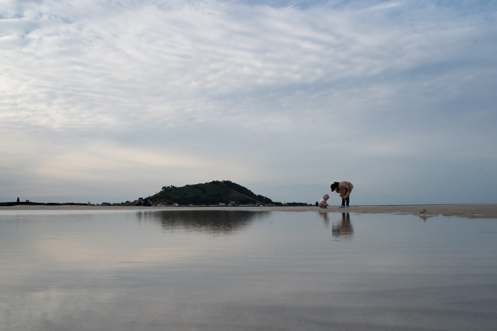 Small figures stand along a shoreline beneath a broad pale sky.