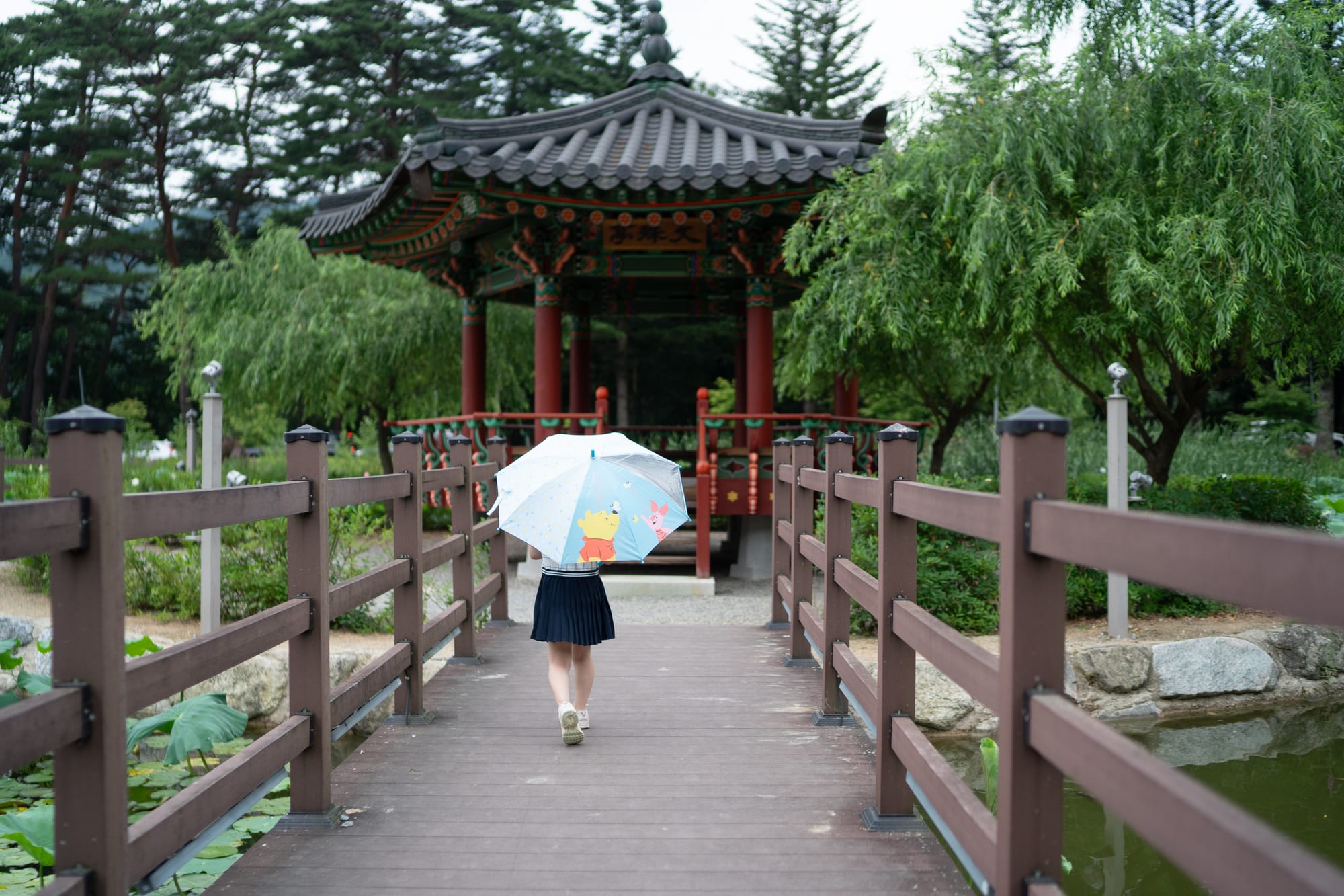 A child walks beneath an umbrella toward a pavilion among trees.